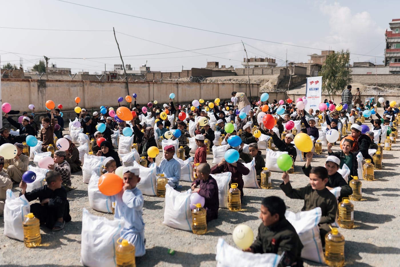 Children seated outdoors with food aid sacks and cooking oil jugs during a community distribution event.