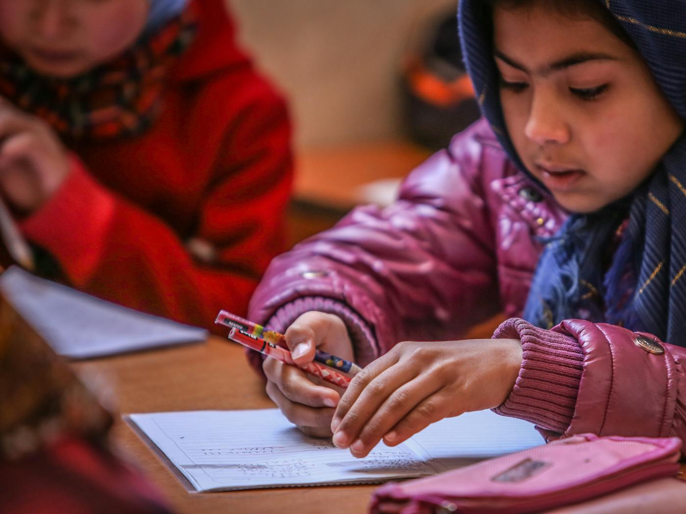 Young student in a headscarf focused on writing at a wooden desk in a classroom, pens and paper in view.