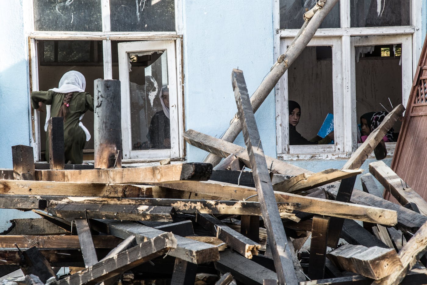 Children at windows of a damaged school building with debris in the foreground — learning amid crisis.